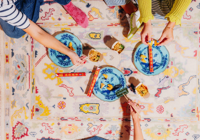 Two people sitting on a colorful rug with plates of food and books.