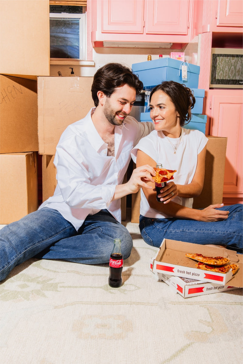 Man and woman sitting on the floor with pizza boxes and a bottle of Coca-Cola in a room with pink cabinets.